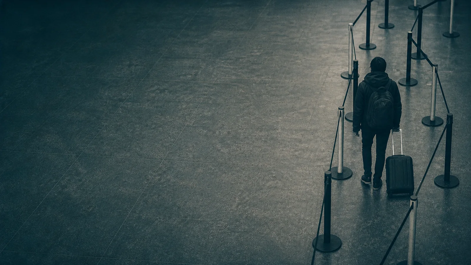 An anonymous traveler with a suitcase stands inside a narrowing airport queue on a dark teal floor.